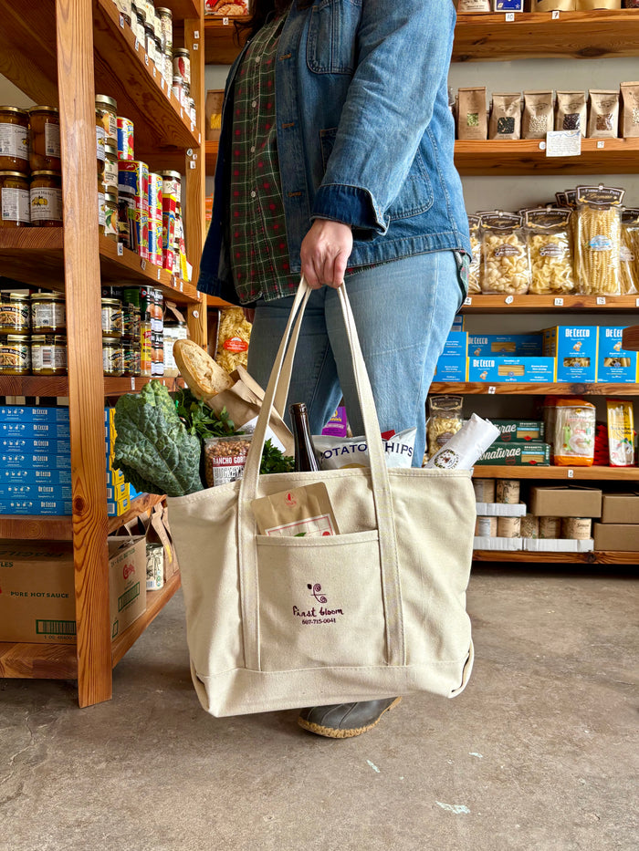 A person holding a beige canvas market tote with two handles and a front slip pocket, standing in a store.