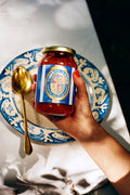 Hand holding a jar of tomato sauce with a decorative label on a table with a blue and white plate and gold spoon.