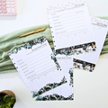 A styled flat-lay of several printed budget and savings goal tracker sheets with floral greenery borders, arranged on a white surface with a folded sage green cloth, a gold pen, a small plant, and part of a pink calculator visible. Disc Bound Planner. Discbound