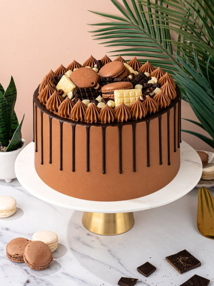 Whole chocolate celebration cake on a gold cake stand with macarons scattered on the marble