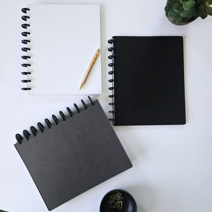 Three discbound leather covers in white, black, and dark gray are arranged on a white desk with a gold crystal pen, a small succulent, and a black dish.