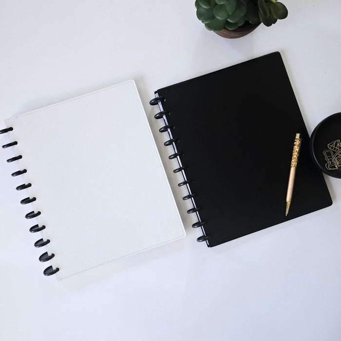 Two disc bound notebooks or planners on a white surface, one white with black spiral binding and one black with black spiral binding. A gold crystal pen and a small black dish with gold geometric designs rest on the black discbound notebook, with a small potted succulent in the top corner.