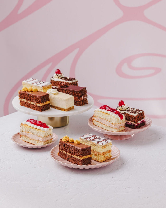 Selection of cake slices on pink plates and a gold cake stand, including cheesecake and Black Forest