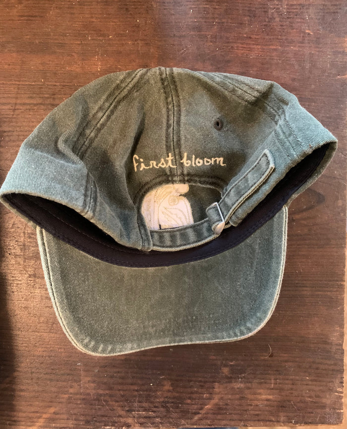 Back view shot of green baseball hat with first bloom logo text on the back, displayed on a wooden surface.