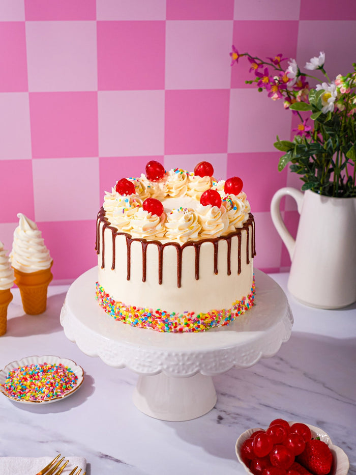 Whole Sweet Treat Sundae Cake topped with cream swirls and glace cherries on a white stand