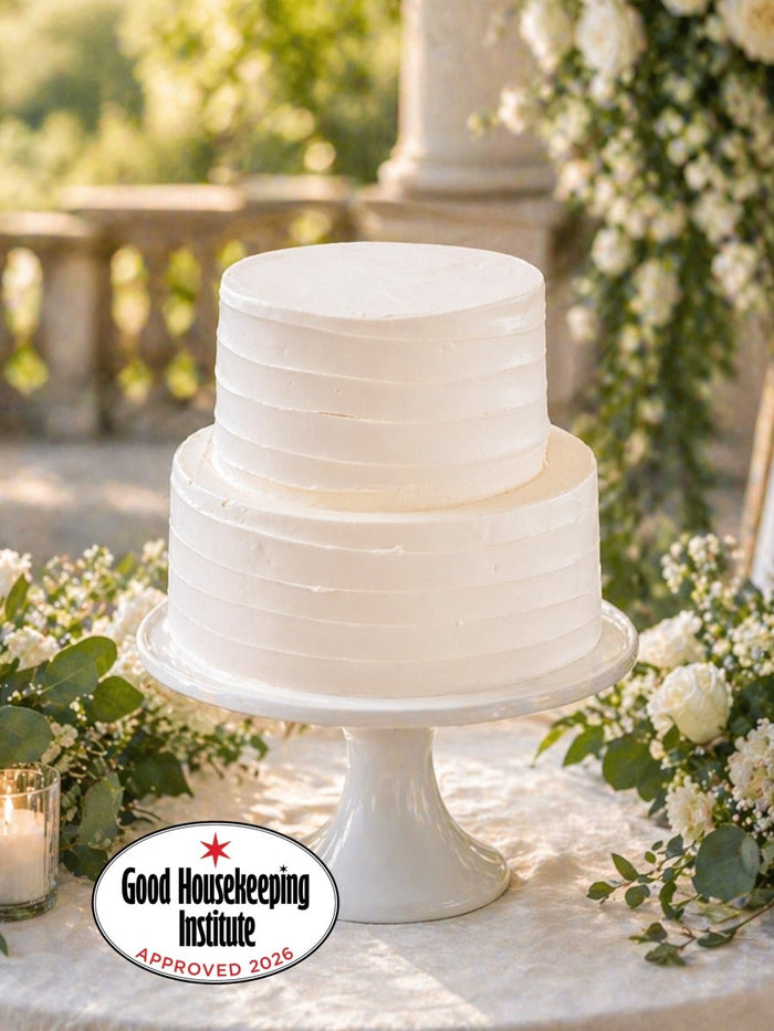 Two tier vanilla wedding cake in smooth buttercream on a white stand, surrounded by foliage